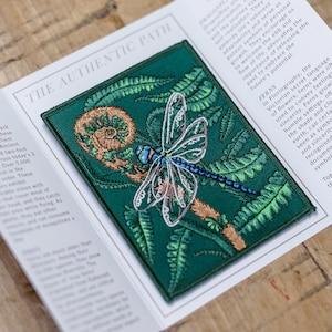May include: An embroidered patch featuring a dragonfly with blue wings and a fern frond. The patch is surrounded by a green border.