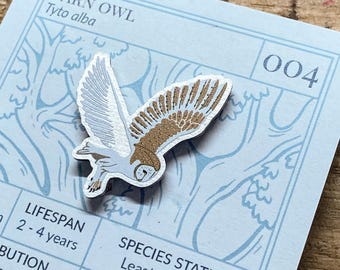 May include: A wooden barn owl pin with a white and brown design. The pin is on a blue card with the text "Barn Owl Tyto alba" and "004". The card also includes information about the owl's lifespan, distribution, and species status.