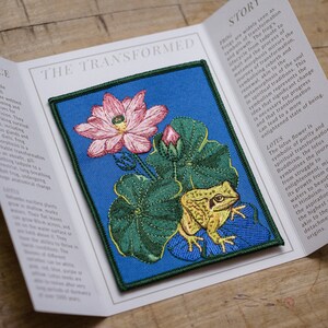 May include: Embroidered patch with a pink lotus flower, a frog, and lily pads on a blue background. The patch has a green border. The text "THE TRANSFORMED" is visible.