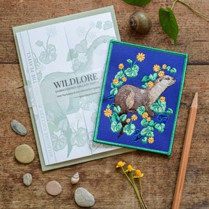 May include: A blue embroidered patch featuring a brown otter surrounded by yellow flowers and green leaves. The patch has a green border and the text "WILDLORE EMBROIDERED GALLERY PATCH" is printed at the top.