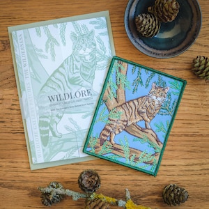May include: An embroidered patch depicting a wildcat on a tree, set against a light blue backdrop, with a green border. A card with the text "WILDLORE" and a wildcat illustration is also visible. The scene is set on a wooden surface.