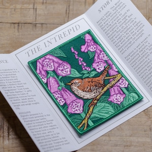 May include: Embroidered patch with a brown wren on a branch, surrounded by purple foxglove flowers on a green backdrop. The patch has a green border and the text "THE INTREPID".
