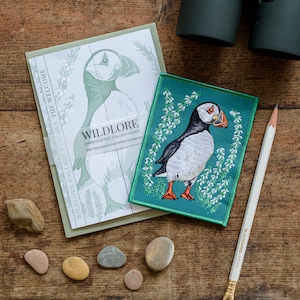 Könnte beinhalten: Ein grüner gestickter Aufnäher mit einem weißen und orangefarbenen Papageitaucher auf grünem Hintergrund. Der Aufnäher trägt den Text "WILDLORE EMBROIDERED GALLERY PATCH" und "With the Science & Story Behind Each Species".