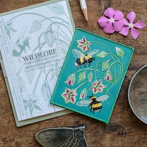 May include: A teal blue embroidered patch featuring two bumblebees and white flowers with pink centres. The patch has the text "WILDLORE EMBROIDERED GALLERY PATCH" and "With The Science & Story Behind Each Species".