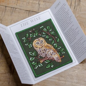 May include: An embroidered patch on a green background, depicting an owl perched on a branch with green leaves. The text "THE WISE" is printed above the owl and "STORY" is printed to the right of the owl. The text describes the owl as a symbol of wisdom and foresight.