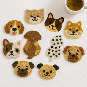 Puede incluir: Una colección de posavasos hechos a mano con forma de perro, en varias razas y colores. Los posavasos presentan expresiones faciales y patrones detallados, incluyendo un dálmata, un carlino y un beagle. Una taza de té está en el fondo.