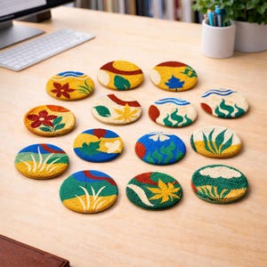 May include: A collection of twelve round, embroidered coasters with various nature-inspired designs. Each coaster features a different pattern, including flowers, leaves, and abstract shapes, in a palette of yellow, blue, green, and red. The coasters are arranged on a light-colored wooden surface.