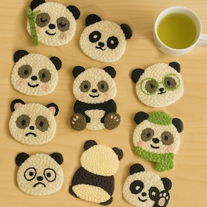 May include: A collection of handmade panda bear coasters in various designs, including one with a bamboo stalk. The coasters are primarily cream-colored with black accents, and some feature pink cheeks and green accessories. A cup of green tea sits nearby.