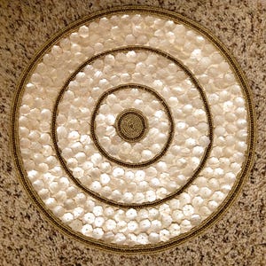 May include: Round, cream-colored placemat with a floral pattern made of small, shell-like pieces. The placemat features concentric circles of dark brown beads and a gold beaded border. The center is a tightly wound spiral of gold beads.