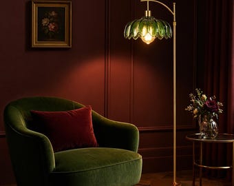 May include: A green glass floor lamp with a scalloped shade and a brass-colored metal frame. The lamp is next to a green velvet armchair with a red pillow. A small table with a vase of flowers is also visible.