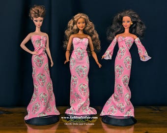 May include: Three Barbie dolls wearing pink and white floral print dresses. The dresses have a fitted bodice and a flowing skirt. The dolls are standing on black stands.