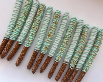May include: Close-up of several chocolate-covered pretzel rods. The pretzels are dipped in white chocolate, drizzled with light blue chocolate, and decorated with gold sprinkles. The image shows a sweet treat.