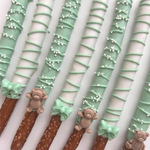 May include: Close-up of decorated pretzel rods. The pretzels are dipped in white and light green icing, with a spiral design and white sprinkles. Each pretzel has a small teddy bear and a bow. A sweet treat.