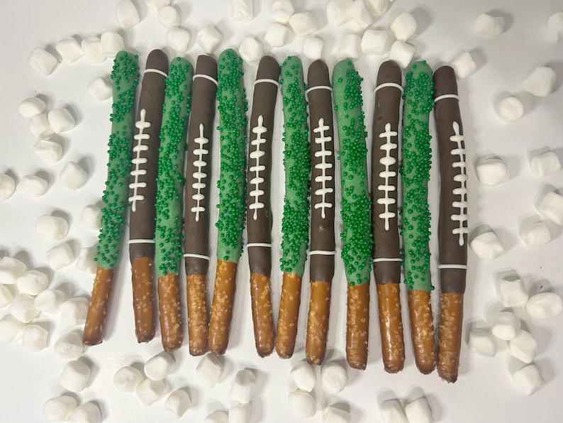 May include: Pretzel rods dipped in chocolate and decorated to resemble footballs. Some are covered in green sprinkles, others have white laces. Marshmallows surround the treats on a white surface.