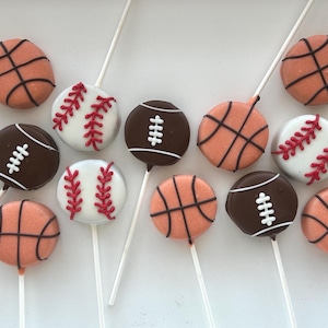 May include: A collection of cake pops decorated to resemble sports balls. The pops include basketballs, baseballs with red stitching, and footballs with white stripes. The pops are on white sticks, perfect for a sports-themed party.