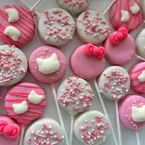 May include: An assortment of pink and white cake pops on white sticks. Some are decorated with pink sprinkles, others with pink bows, white cat-shaped candies, and pink stripes. A sweet treat.