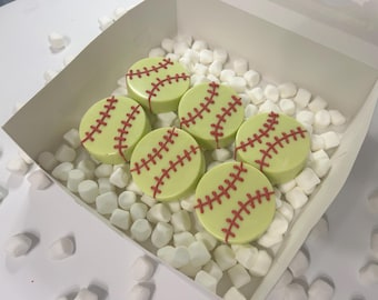 Op de afbeelding: Zes groene en witte softball-vormige chocolade traktaties met rode stiksels zijn gerangschikt in een witte doos gevuld met witte marshmallows.