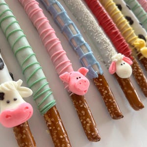 May include: Close-up of decorated pretzel rods with farm animal designs. The pretzels are dipped in white, pink, blue, and red chocolate, with green, pink, and white icing. Each pretzel features a different farm animal face.