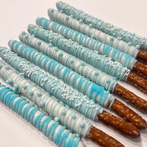 May include: Several chocolate-covered pretzel rods. The pretzels are dipped in light blue and white chocolate, with sprinkles and pearl decorations. The pretzel rods are arranged in a diagonal pattern on a white surface.