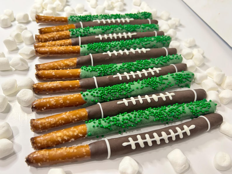 May include: Pretzel rods dipped in chocolate and decorated to resemble footballs. Some are covered in green sprinkles, others have white icing laces. Marshmallows are scattered around the treats.