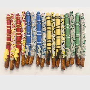 May include: An assortment of chocolate-covered pretzel rods, decorated in various colors. The pretzels are dipped in white chocolate and adorned with sprinkles, stripes, and lightning bolt-shaped candies. The color scheme includes red, yellow, blue, and green.