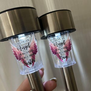 May include: Two solar garden lights with silver tops and stems. Each light has a clear plastic section with the text "I HAVE AN Angel IN HEAVEN CALL HER Sister" and "I HAVE AN Angel IN HEAVEN CALL HER Nan" with pink angel wings.