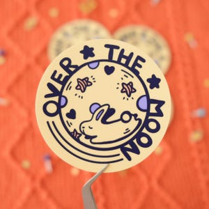May include: A yellow circle sticker with the text "Over the Moon" in a black outline. The moon is depicted as a yellow circle with a purple and blue pattern, stars, and a white bunny sleeping on it.