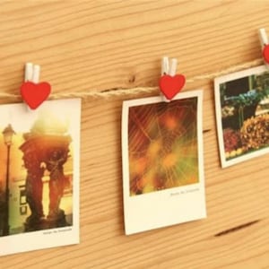 May include: A string of photos is hung on a wall with red heart-shaped clothespins. The photos are of various subjects, including a street scene, a garden, and a close-up of a tree.