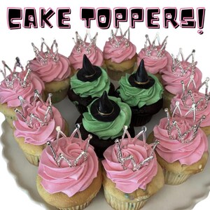 Pode incluir: Uma coleção de cupcakes decorados com glacê rosa e verde. Alguns cupcakes têm coroas prateadas no topo, enquanto outros têm chapéus de bruxa pretos. Os cupcakes estão dispostos em um prato branco. O texto "CAKE TOPPERS!" está escrito em letras rosa na parte superior da imagem.