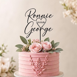 Puede incluir: Un pastel glaseado rosa decorado con rosas rosas, vegetación y un diseño de hojas decorativas. Un adorno negro para pastel dice "Ronnie" y "George" con un corazón y la fecha "12.12.26".