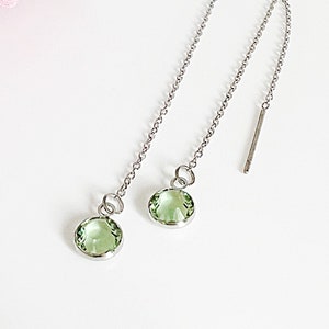 May include: A pair of silver threader earrings with round, light green gemstones. The earrings are displayed on a white surface with a pink flower in the background. The gemstones are set in a silver bezel.