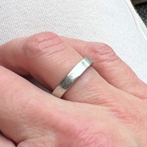 May include: A silver ring with a flat, rectangular band is worn on a finger. The ring has a slightly hammered texture, reflecting light. The ring is a simple, minimalist design.