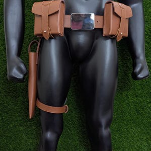 May include: A brown leather belt with two pouches on either side and a silver buckle. The belt is worn on a mannequin with brown leather leg straps and pouches.
