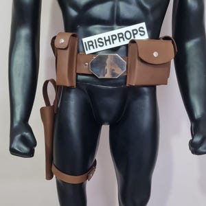 Puede incluir: Un cinturón utilitario de cuero marrón con dos bolsillos, una funda y una hebilla plateada. El cinturón se muestra en un maniquí negro. El texto "IRISHPROPS" es visible en una etiqueta blanca.