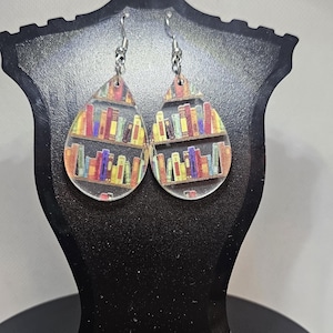 Könnte beinhalten: Ein Paar tropfenförmige Ohrringe mit einem transparenten Hintergrund und einem bunten Bild von Büchern in einem Bücherregal. Die Ohrringe sind silberfarben und haben einen Hakenverschluss.