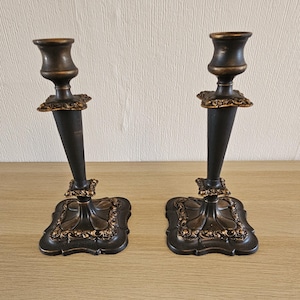 Op de afbeelding: Twee antieke kandelaars in donker brons met gouden details. Elke houder heeft een vierkante basis met sierlijke patronen, een donkere steel en een kopvormige top. Geschikt voor standaard kaarsen.