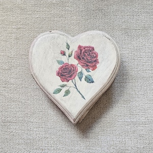 Peut inclure: Une plaque en bois en forme de cœur blanche avec un motif peint de deux roses rouges et de feuilles vertes.