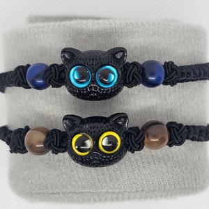 Könnte beinhalten: Zwei schwarze elastische Armbänder mit schwarzen Katzenanhängern. Das obere Armband hat blaue Perlen und das untere Armband hat braune Perlen.