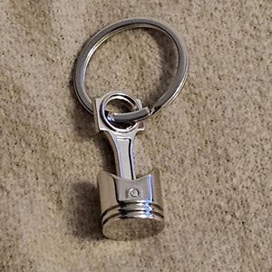 Può includere: Un portachiavi in metallo argentato con un modello dettagliato di un pistone. Il portachiavi ha un anello argentato per attaccare le chiavi.