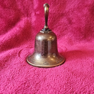 K&ouml;nnte beinhalten: Eine kleine, bronzefarbene Glocke mit gebogenem Griff. Die Glocke hat einen ausgestellten Sockel mit ge&auml;tztem Text und herzf&ouml;rmigen Designs. Sie liegt auf einer strukturierten, rosa Oberfl&auml;che.