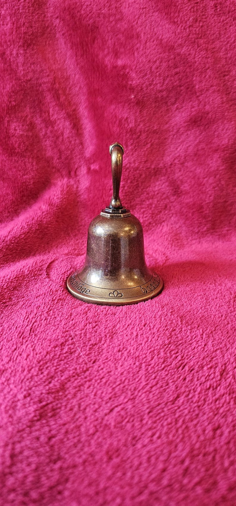 K&ouml;nnte beinhalten: Eine kleine, bronzefarbene Glocke mit gebogenem Griff. Die Glocke hat einen ausgestellten Sockel mit ge&auml;tztem Text und herzf&ouml;rmigen Designs. Sie liegt auf einer strukturierten, rosa Oberfl&auml;che.