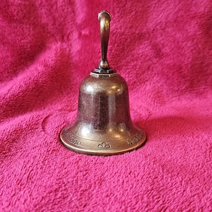 K&ouml;nnte beinhalten: Eine bronzefarbene Glocke mit gebogenem Griff. Die Glocke hat einen ausgestellten Sockel mit dekorativen Elementen und Text. Die Glocke befindet sich vor einem rosa, strukturierten Hintergrund.