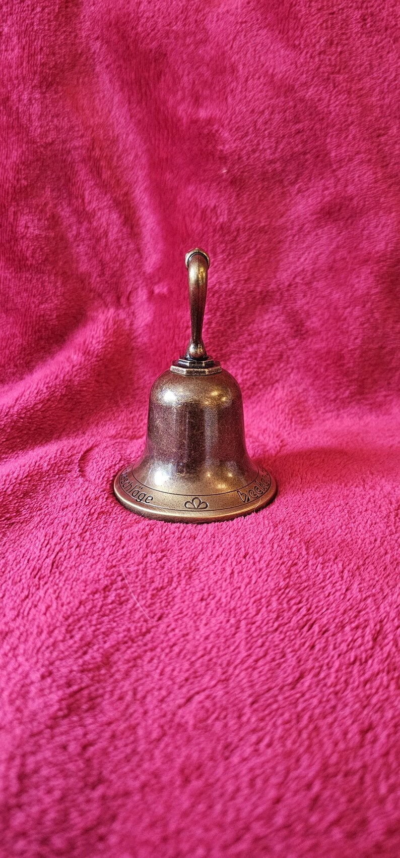 K&ouml;nnte beinhalten: Eine bronzefarbene Glocke mit gebogenem Griff. Die Glocke hat einen ausgestellten Sockel mit dekorativen Elementen und Text. Die Glocke befindet sich vor einem rosa, strukturierten Hintergrund.