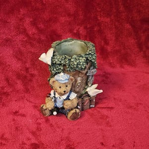 Könnte beinhalten: Ein dekorativer Pflanztopf mit einem Teddybären mit blauer Mütze und Latzhose, der neben einem Baumstamm sitzt. Der Baum hat eine grüne, belaubte Spitze und einen kleinen Koffer. Zwei weiße Tauben sitzen auf dem Baum.