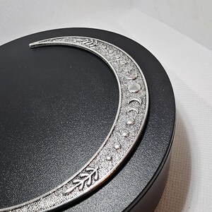 Puede incluir: Un amuleto de plata en forma de media luna con un dise&ntilde;o detallado de hojas y puntos. El amuleto est&aacute; en un expositor circular negro.