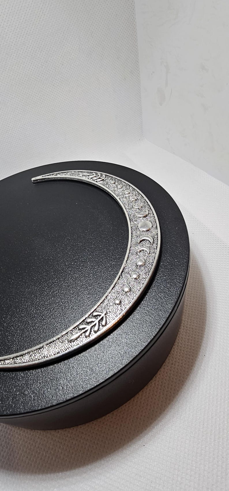 Puede incluir: Un amuleto de plata en forma de media luna con un dise&ntilde;o detallado de hojas y puntos. El amuleto est&aacute; en un expositor circular negro.