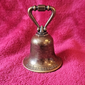 K&ouml;nnte beinhalten: Bronzefarbene Glocke mit Griff, auf rosa Untergrund. Die Glocke hat einen ausgestellten Sockel mit dem Text "Becker-beschl&auml;ge". Der Griff ist gew&ouml;lbt und an der Oberseite der Glocke befestigt.
