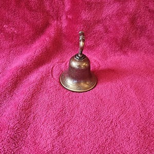 K&ouml;nnte beinhalten: Eine kleine Glocke im antiken Stil mit Bronze-Finish. Die Glocke hat einen abgerundeten K&ouml;rper und einen gebogenen Griff. Die Glocke steht auf einer rosa, strukturierten Oberfl&auml;che.