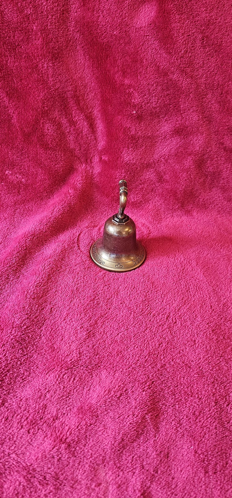K&ouml;nnte beinhalten: Eine kleine Glocke im antiken Stil mit Bronze-Finish. Die Glocke hat einen abgerundeten K&ouml;rper und einen gebogenen Griff. Die Glocke steht auf einer rosa, strukturierten Oberfl&auml;che.