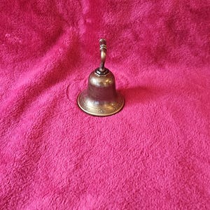 K&ouml;nnte beinhalten: Eine kleine Glocke aus Messing im antiken Stil mit gebogenem Griff und ausgestelltem Sockel. Die Glocke hat eine angelaufene Oberfl&auml;che und ist vor einem rosa Hintergrund platziert. Der Sockel der Glocke hat dekorative Details.